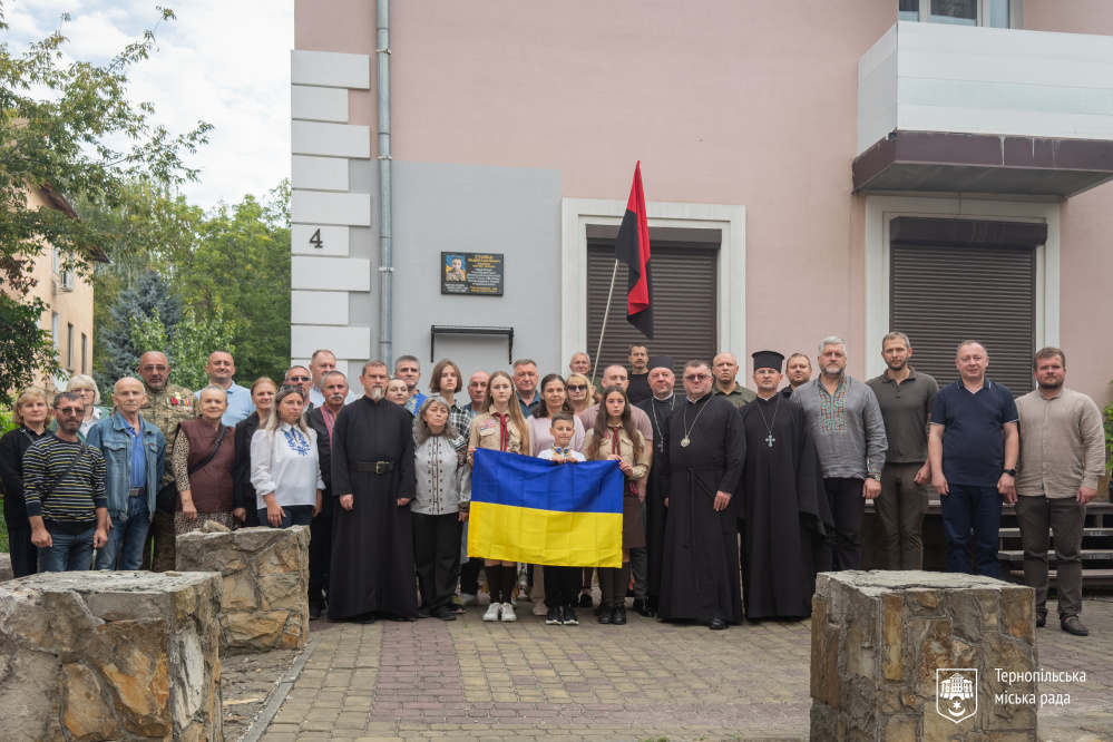 Велика група людей фотографується біля меморіальної дошки. У центрі кілька людей тримають синьо-жовтий прапор України.